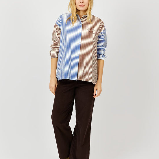 A woman modeling a long-sleeve button-up shirt with a split design: blue and white vertical stripes on one side and beige and brown vertical stripes on the other, paired with dark brown wide-leg pants and patterned shoes, against a light gray studio background.