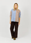 A woman modeling a long-sleeve button-up shirt with a split design: blue and white vertical stripes on one side and beige and brown vertical stripes on the other, paired with dark brown wide-leg pants and patterned shoes, against a light gray studio background.