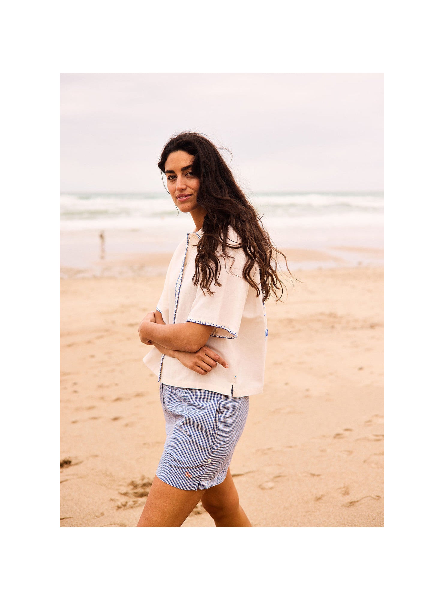 Woman standing on a beach wearing a light-colored jacket and blue shorts.
